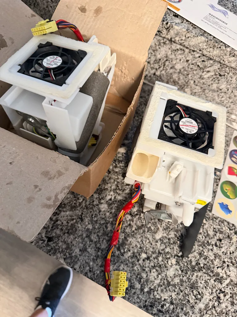 Two white plastic assemblies with black fans and wiring harnesses side by side on a granite counter, one of them in a cardboard box